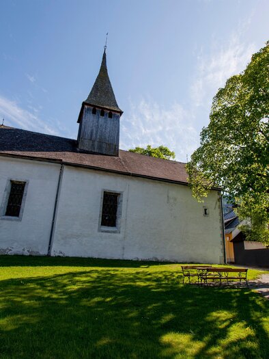 An old church with a pointed tower and a large tree in the foreground. The lawn is green and the surroundings feel calm and inviting.
