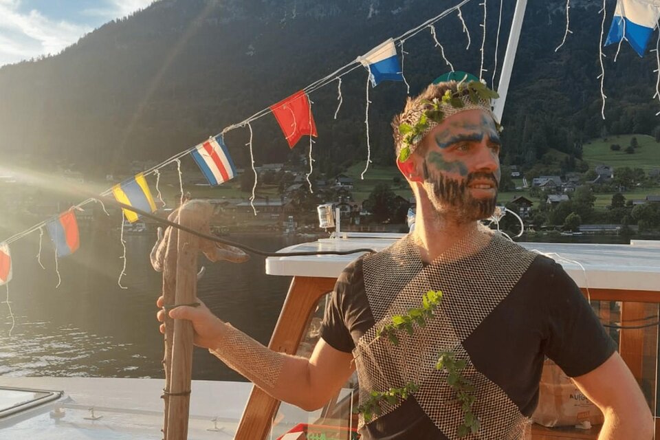 Ein Mann in etwas traditioneller Kleidung steht auf einem Boot. Er hält einen Stock und trägt eine Blumenkrone sowie Gesichtsbemalung. | © Schifffahrt Grundlsee Toplitzsee