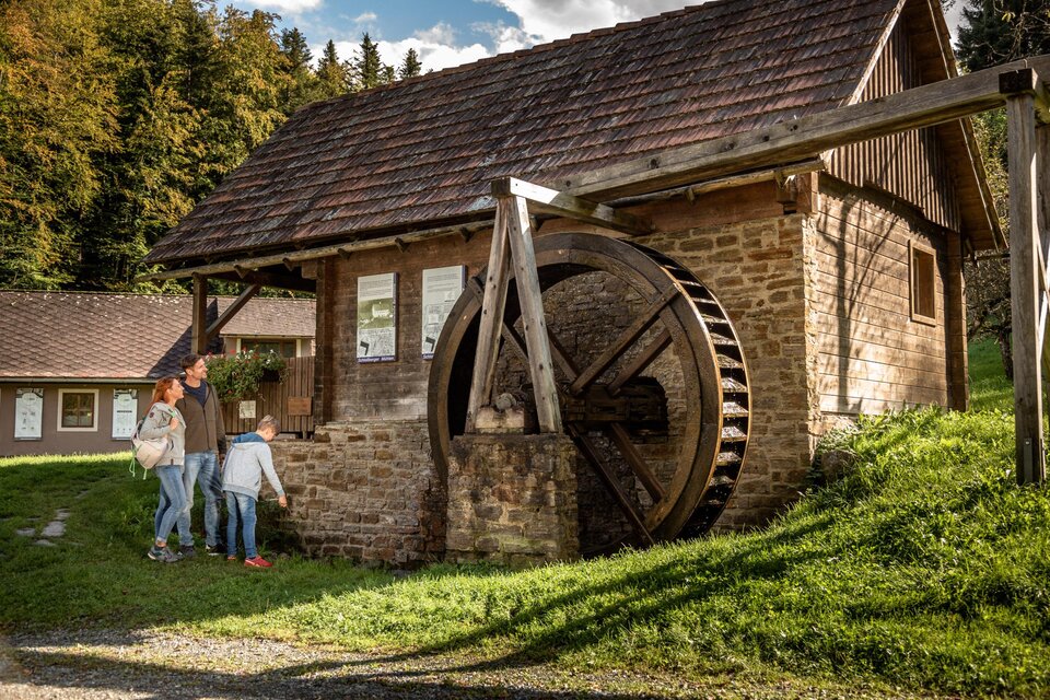 Ein historisches Wasserrad neben einem kleinen Steinhaus. Kinder erkunden die Umgebung auf einer grünen Wiese. | © Stephan Friesinger