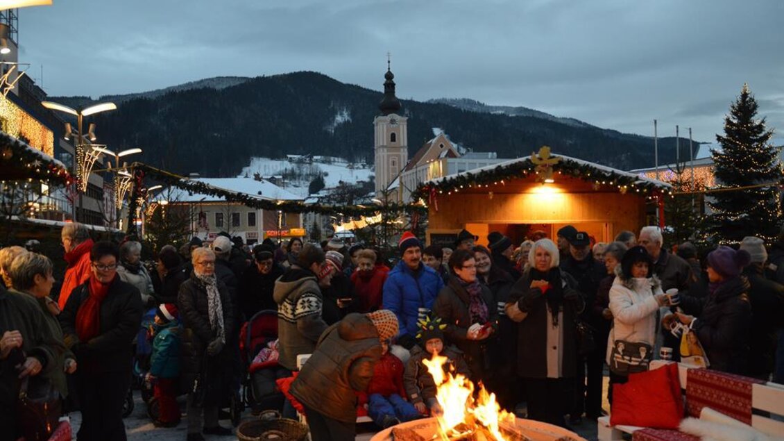 Ein belebter Weihnachtsmarkt mit vielen Menschen und festlicher Beleuchtung. In der Mitte brennt ein Lagerfeuer, während im Hintergrund eine Kirche zu sehen ist. | © Mürzzuschlag Agentu, Baumann
