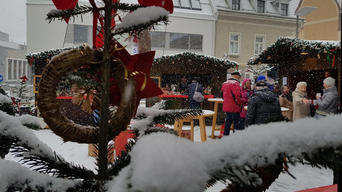 Ein stimmungsvolles Weihnachtsmarkt-Szenario im Schnee. Menschen stehen um festlich dekorierte Buden herum, während Schneeflocken sanft fallen. | © Stadt Mürzzuschlag, Bettina Rinnhofer