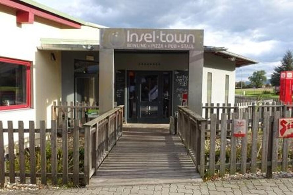 Ein Eingangsbereich zu "Insel-town" mit einer Holzbrücke und einem modernen Gebäude. Es bietet Bowling, Pizza und eine Bar. | © Inseltown