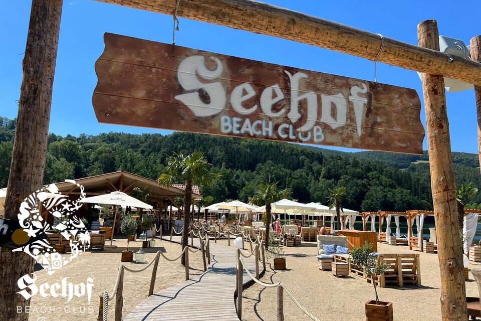 Ein einladender Beach Club mit einem Holzschild, das "See-hof Beach Club" zeigt. Im Hintergrund sind Liegen, Sonnenschirme und eine grüne Umgebung zu sehen. | © Seehof-Beachclub