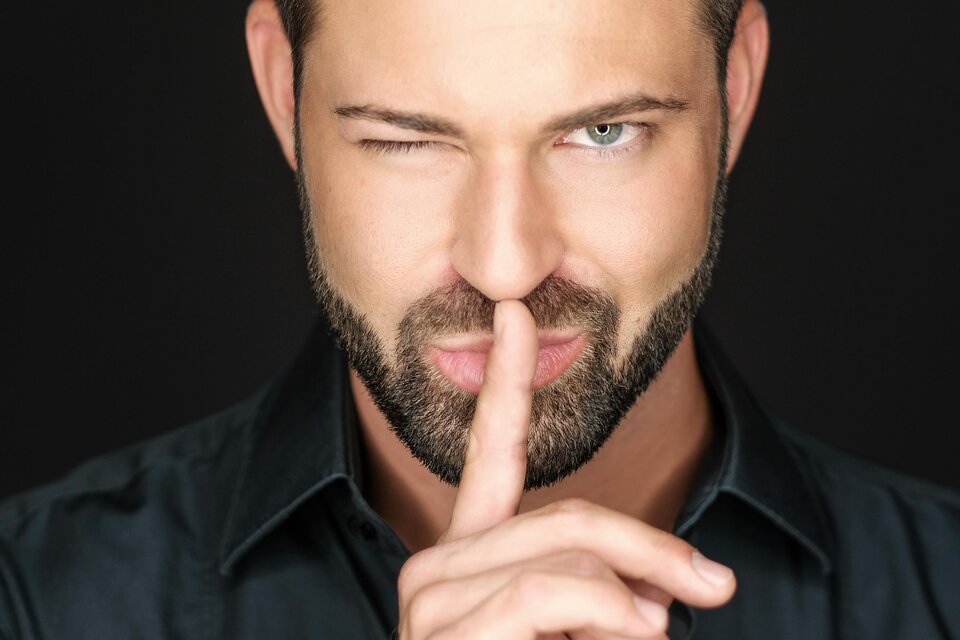 Ein Mann mit Bart und witzigem Blick hält seinen Zeigefinger an die Lippen. Er trägt ein schwarzes Hemd und hat einen geheimnisvollen Ausdruck. | © Jean Van Luelik