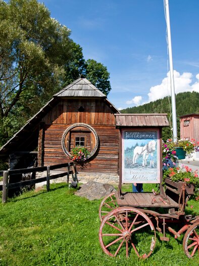 A rustic wooden building with a simple roof and a round window. In front stands an old wooden wagon wheel and blooming plants adorn the surroundings.