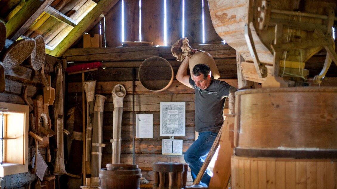 Ein Mann arbeitet in einer traditionellen Mühle. Im Hintergrund sind verschiedene Holzwerkzeuge und Maschinen zu sehen.