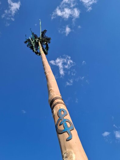 A tall pole with a braided crown on top and a decorative symbol on the side. The sky is blue with a few white clouds. | © Musikverein Bretstein
