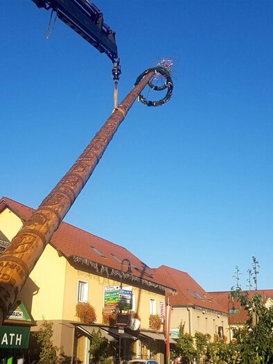 Ein hoher Kranausleger, der eine kunstvolle Figur in die Luft hebt. Im Hintergrund sieht man elegante, traditionelle Gebäude unter einem klaren blauen Himmel. | © FF Gamlitz
