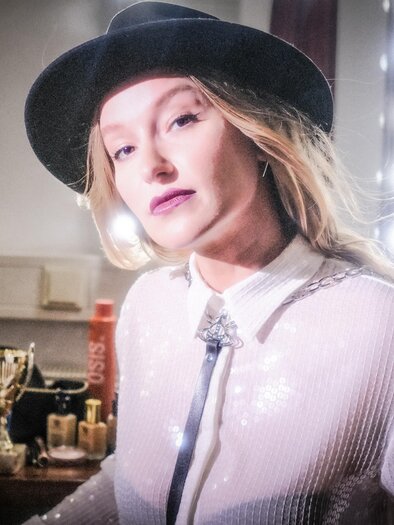 The pretty cabaret artist Malarina presents herself in an artist's dressing room in front of the lighted makeup mirror wearing a hat, white blouse, cheeky black skirt, fishnet stockings, and black boots. | © Oststeiermark Tourismus