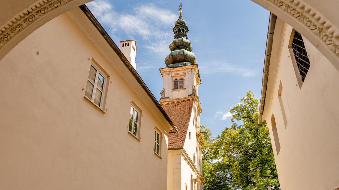 Ein malerischer Durchgang führt zu einem historischen Gebäude mit einem hohen Turm. Umgeben von grünen Bäumen und einer schönen Umgebung spazieren Menschen entlang des Weges. | © Wolfgang Spekner