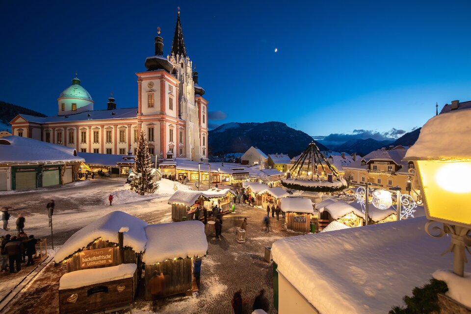 Ein malerischer Weihnachtsmarkt in einer verschneiten Stadt. Im Hintergrund ist eine beeindruckende Kirche und ein beleuchteter Weihnachtsbaum zu sehen. | © TV Hochsteiermark