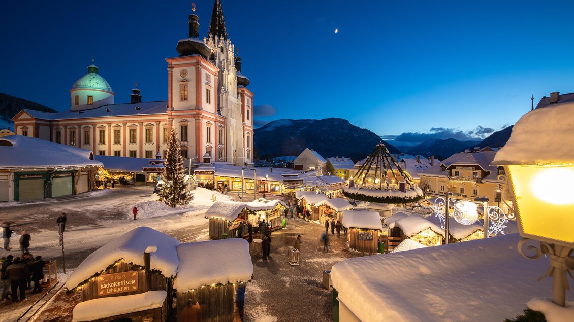 Ein malerischer Weihnachtsmarkt in einer verschneiten Stadt. Im Hintergrund ist eine beeindruckende Kirche und ein beleuchteter Weihnachtsbaum zu sehen. | © TV Hochsteiermark