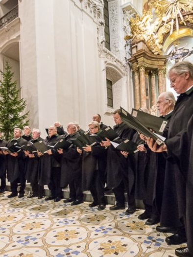 A choir of men in long, dark robes sings in a church. In the background, a Christmas tree and elaborate architecture can be seen. | © Fred Lindmoser