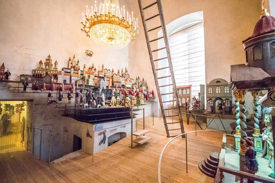 Ein historisches Museum mit vielen kunstvoll gestalteten Figuren und Modellen. Eine Leiter führt zu einem höheren Bereich, und ein Kronleuchter beleuchtet den Raum. | ©  Fred Lindmoser