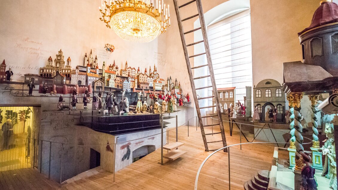 Ein historisches Museum mit vielen kunstvoll gestalteten Figuren und Modellen. Eine Leiter führt zu einem höheren Bereich, und ein Kronleuchter beleuchtet den Raum. | ©  Fred Lindmoser