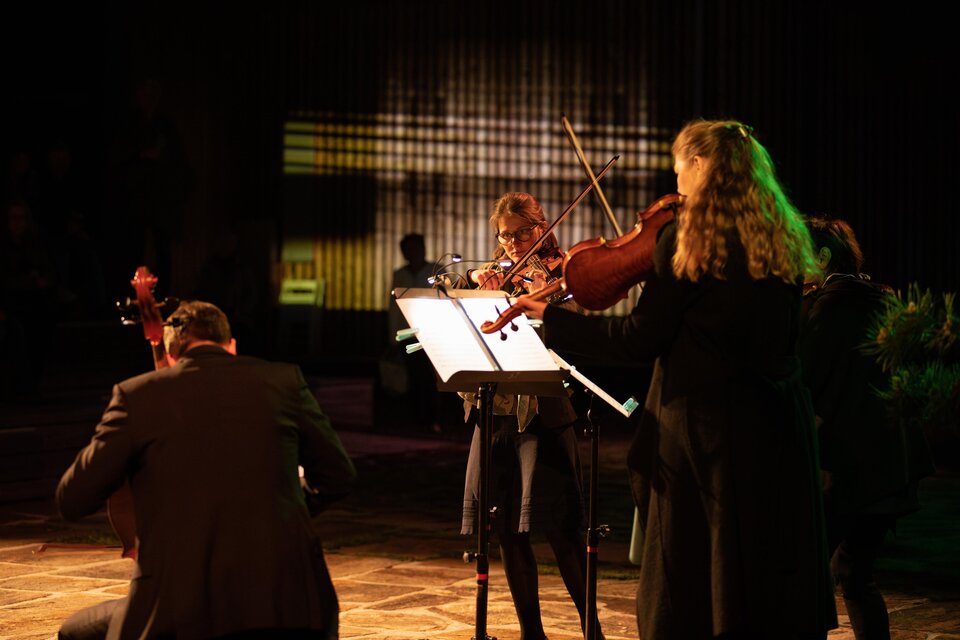 Eine Gruppe von Musikern spielt auf einer Bühne. Sie sind mit verschiedenen Streichinstrumenten ausgestattet und stehen im Schein von farbigem Licht. | © TVB Südsteiermark – Achromatic Photography