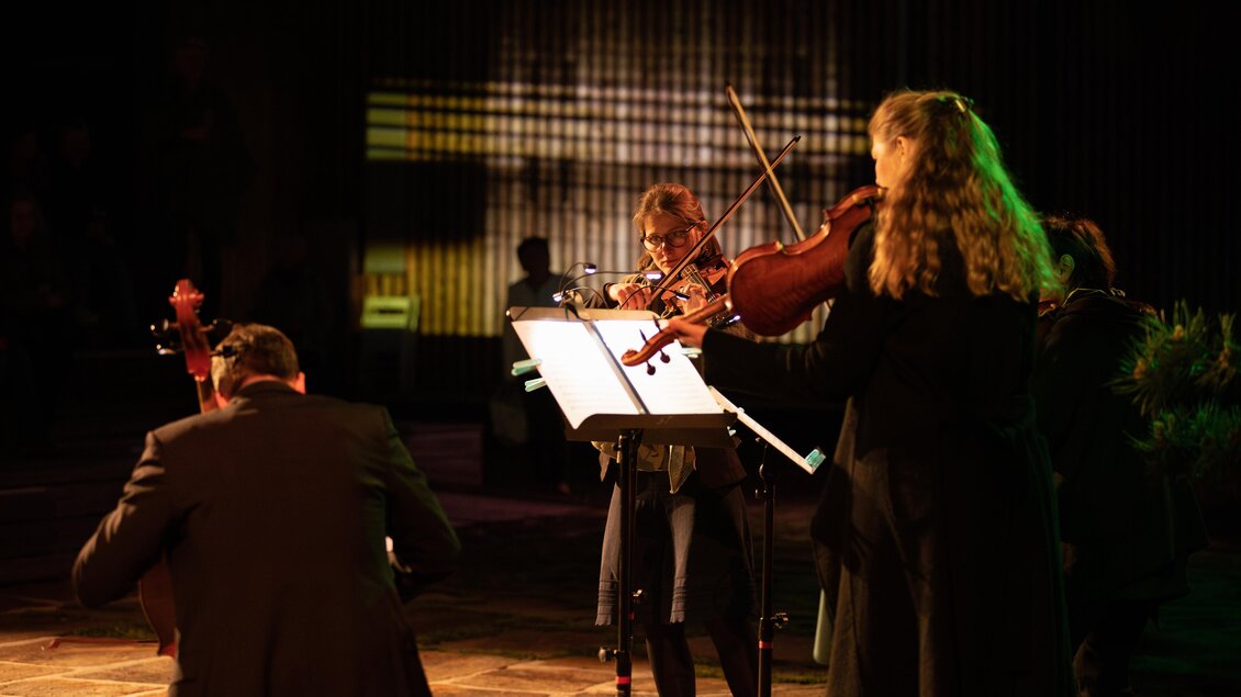 Eine Gruppe von Musikern spielt auf einer Bühne. Sie sind mit verschiedenen Streichinstrumenten ausgestattet und stehen im Schein von farbigem Licht. | © TVB Südsteiermark – Achromatic Photography