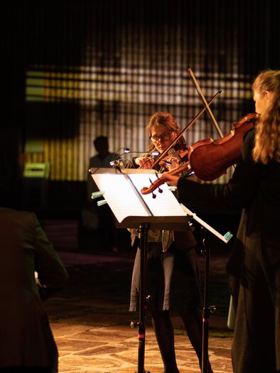 Eine Gruppe von Musikern spielt auf einer Bühne. Sie sind mit verschiedenen Streichinstrumenten ausgestattet und stehen im Schein von farbigem Licht. | © TVB Südsteiermark – Achromatic Photography
