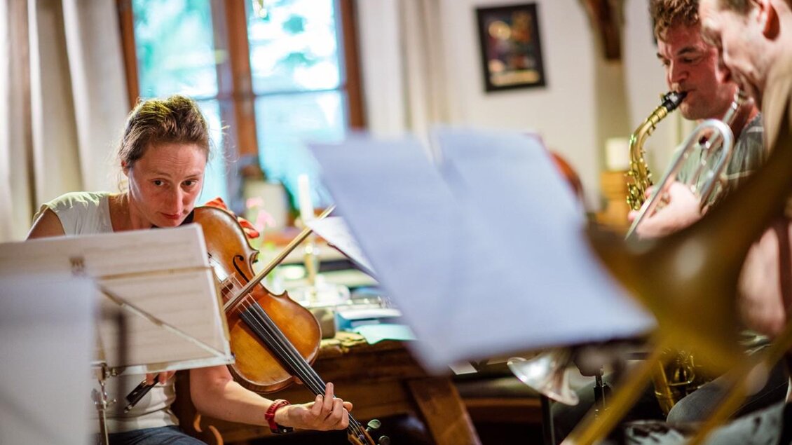 Eine Musikergruppe spielt in einem gemütlichen Raum. Im Vordergrund spielt eine Frau mit einer Violine. | © @Greith Haus