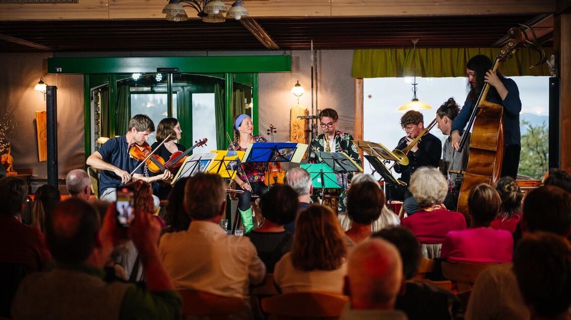 Eine Gruppe von Musikern spielt live auf der Bühne in einem gemütlichen Veranstaltungsort. Das Publikum sitzt aufmerksam und genießt die Darbietung. | © @ Greith Haus