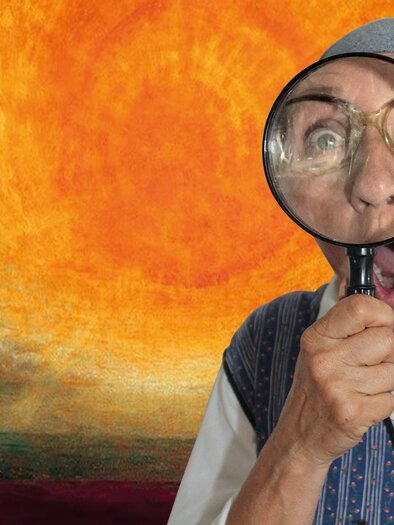 Humorous staging of a woman with a headscarf, curiously looking into the camera with a large magnifying glass. The colorful background and the surprised facial expression give the motif a cheerful, cabaret-like character. To be seen in Straden. | © Astrid Reichhold