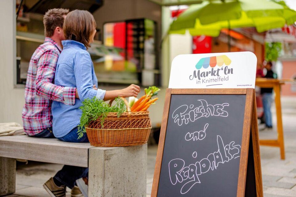 Ein Paar sitzt auf einer Bank und genießt die Zeit zusammen. Im Vordergrund steht ein Schild mit der Aufschrift „Frisches und Regionales“. | © Markt´ln Knittelfeld