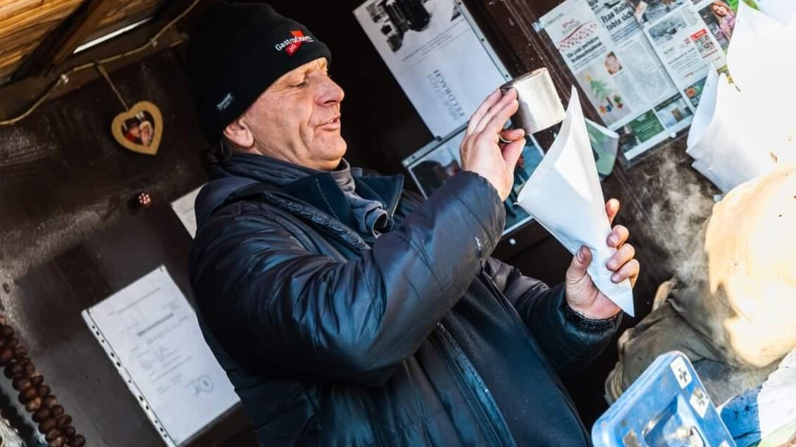 Ein Mann steht vor einem Stand und hält eine Tüte in der Hand. Er lächelt und scheint mit einem Kunden zu interagieren. | © Stadtgemeinde Feldbach