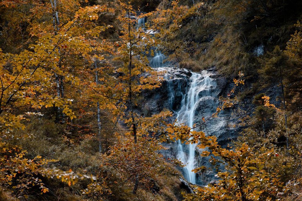 Ein kleiner Wasserfall fließt durch eine herbstliche Landschaft mit orangefarbenen und gelben Blättern. Die Umgebung ist ruhig und naturnah. | © TV Gesäuse
