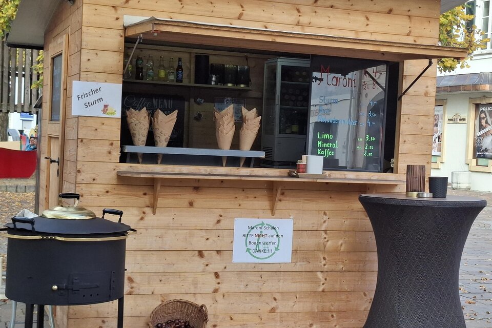 Ein Holzstand mit einem Verkaufsfenster und einer Auswahl an Waffeln. Neben dem Stand steht ein runder Tisch mit einer Schüssel und einem Kochtopf. | © Gerlinde Rath