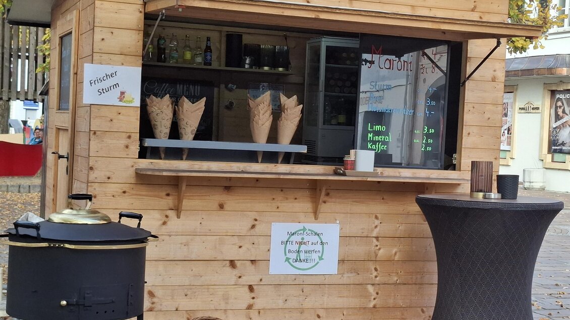 Ein Holzstand mit einem Verkaufsfenster und einer Auswahl an Waffeln. Neben dem Stand steht ein runder Tisch mit einer Schüssel und einem Kochtopf. | © Gerlinde Rath