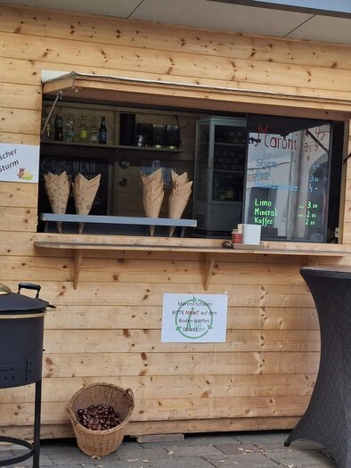 A wooden stall with a sales window and a selection of waffles. Next to the stall, there is a round table with a bowl and a cooking pot. | © Gerlinde Rath