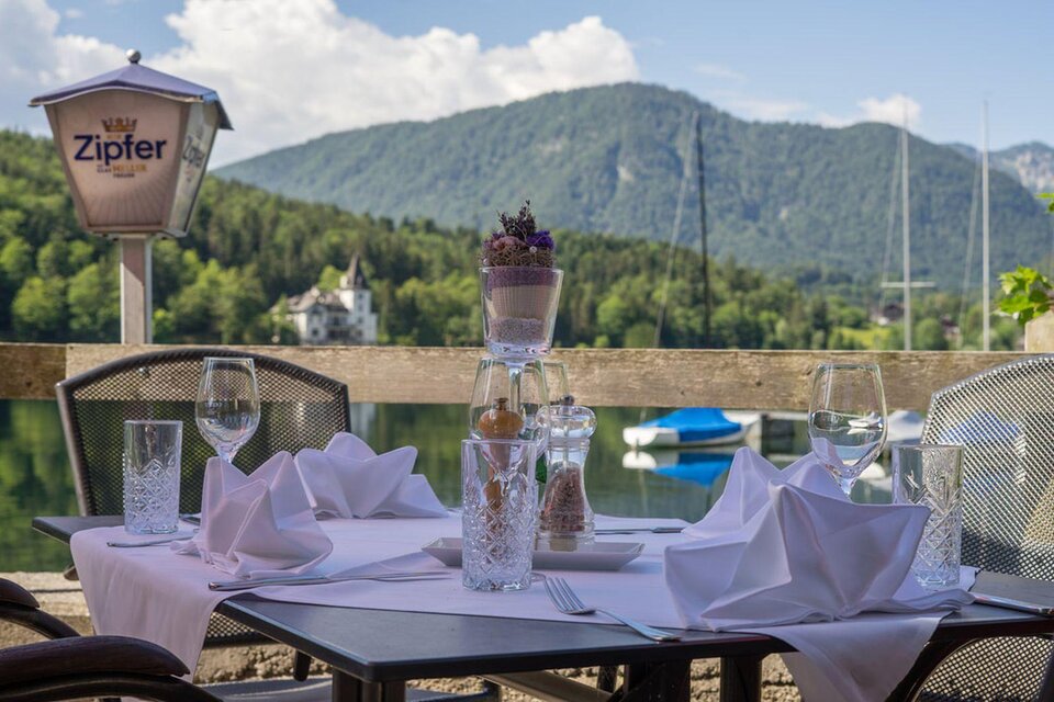 Ein elegant gedeckter Tisch im Dorfwirtshaus Stöckl mit Blick auf den wunderschönen Grundlsee und die umliegenden Berge des Ausseerlandes. Die Tischdekoration umfasst Gläser und Servietten, die schön angeordnet sind. | © Dorfwirtshaus Stöckl