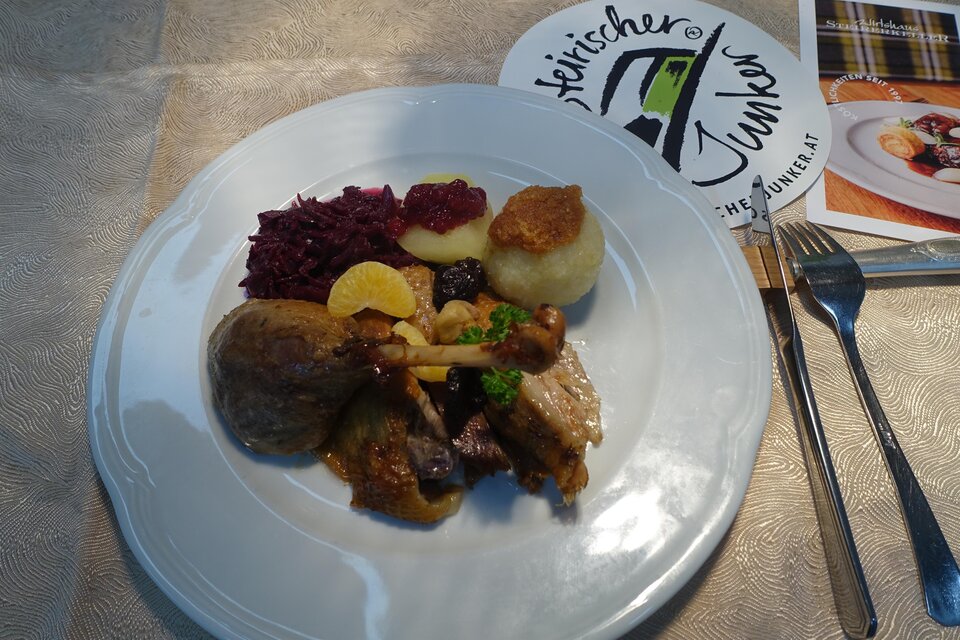 Ein Teller mit verschiedenen Gerichten, einschließlich Fleisch, Knödeln, Rotkraut und einer Fruchtgarni. Im Hintergrund ist eine Speisekartenanordnung zu sehen. | © Kröll