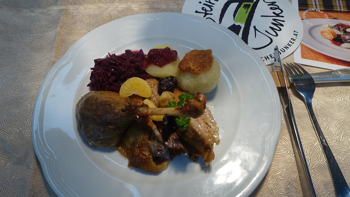 Ein Teller mit verschiedenen Gerichten, einschließlich Fleisch, Knödeln, Rotkraut und einer Fruchtgarni. Im Hintergrund ist eine Speisekartenanordnung zu sehen. | © Kröll