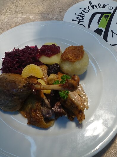 Ein Teller mit verschiedenen Gerichten, einschließlich Fleisch, Knödeln, Rotkraut und einer Fruchtgarni. Im Hintergrund ist eine Speisekartenanordnung zu sehen. | © Kröll