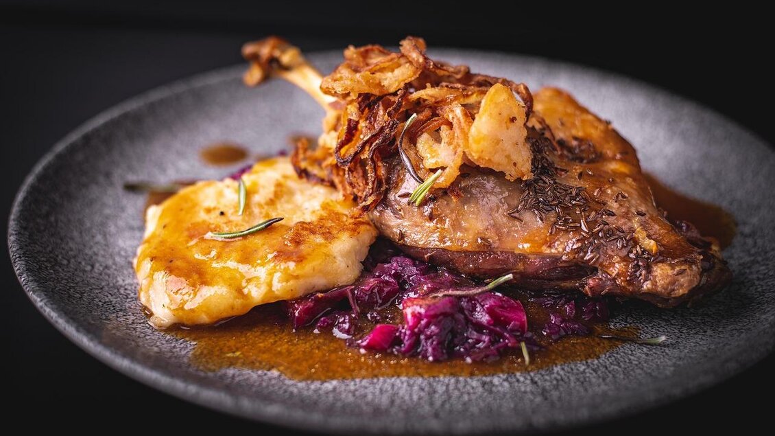 Ein geschmackvoll angerichteter Teller mit Entenkeule, serviert auf Rotkohl und Kartoffelstock. Das Gericht ist mit knusprigen Zwiebeln garniert und sieht sehr appetitlich aus. | © Wirtshaus Steirerkeller