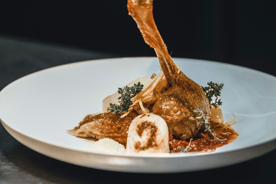 Ein kunstvoll angerichteter Teller mit einem gebratenen Hähnchenbein, umgeben von einer würzigen Sauce. Dazu befinden sich Gemüse und Kräuter als Garnitur. | © Vulkanlandhotel Legenstein
