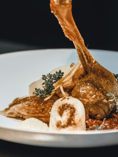 An artistically arranged plate with a roasted chicken leg, surrounded by a spicy sauce. Accompanied by vegetables and herbs as garnish. | © Vulkanlandhotel Legenstein