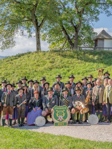 A group of people in traditional clothing poses outdoors. In the background, trees and a small building are visible. | © Musikverein Stainach