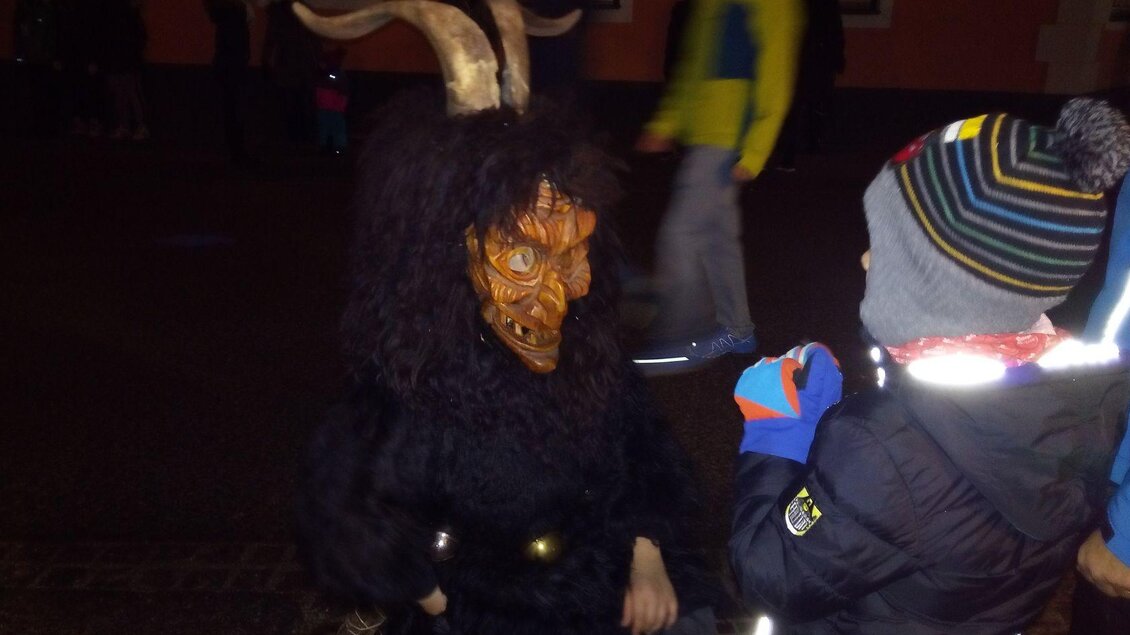 Ein Bild eines Krampus, der vor einem Kind kniet, während das Kind betet. Der Krampus trägt ein schwarzes Fell und eine traditionelle Holzmaske mit zwei Hörnern auf dem Kopf. Die Szene spielt bei Dunkelheit und vermittelt eine mystische, stimmungsvolle Atmosphäre. | © Johanna Provatopoulos