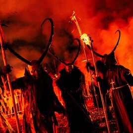 Eine Gruppe von maskierten Figuren mit Geweihen steht in einem feurigen, rauchigen Umfeld. Die düstere Atmosphäre wird durch das orangefarbene Licht intensiviert. | © Gstoder Pass Ranten