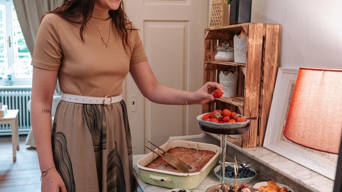 Eine lächelnde Frau steht an einem Tisch mit köstlichen Snacks und Desserts. Im Hintergrund sind eine gemütliche Einrichtung und eine Lampe zu sehen. | © Landgut Riegerbauer