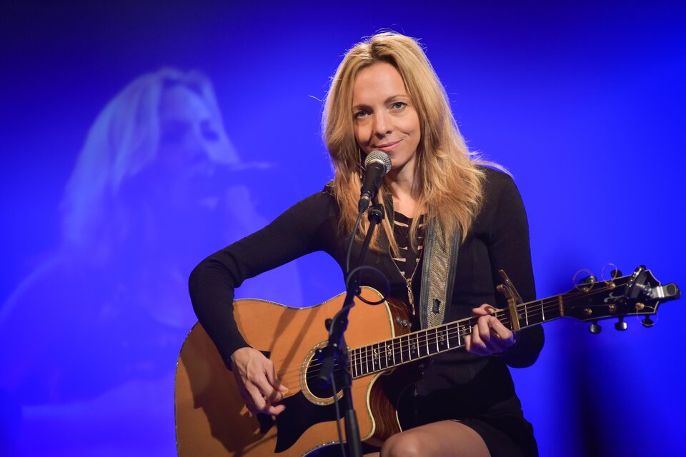 Eine Sängerin mit blonden Haaren spielt Gitarre auf der Bühne. Im Hintergrund ist ein sanftes Licht und eine verschwommene Projektion von ihr zu sehen. | © Christoph Huber