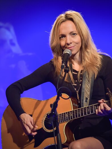 Eine Sängerin mit blonden Haaren spielt Gitarre auf der Bühne. Im Hintergrund ist ein sanftes Licht und eine verschwommene Projektion von ihr zu sehen. | © Christoph Huber