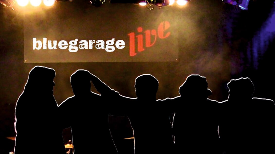 Eine Gruppe von Menschen steht silhouettenartig vor einer Bühne mit bunten Lichtern. Im Hintergrund ist ein Banner mit der Aufschrift "bluegarage live" zu sehen. | © Bluegarage