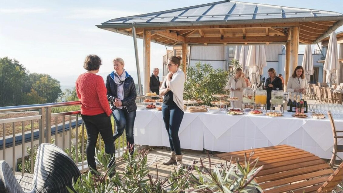 Eine Gruppe von Menschen steht auf einer sonnigen Terrasse mit einem Buffet. Im Hintergrund sind Tische und eine schöne Aussicht zu sehen. | © Hotel Retter und KI