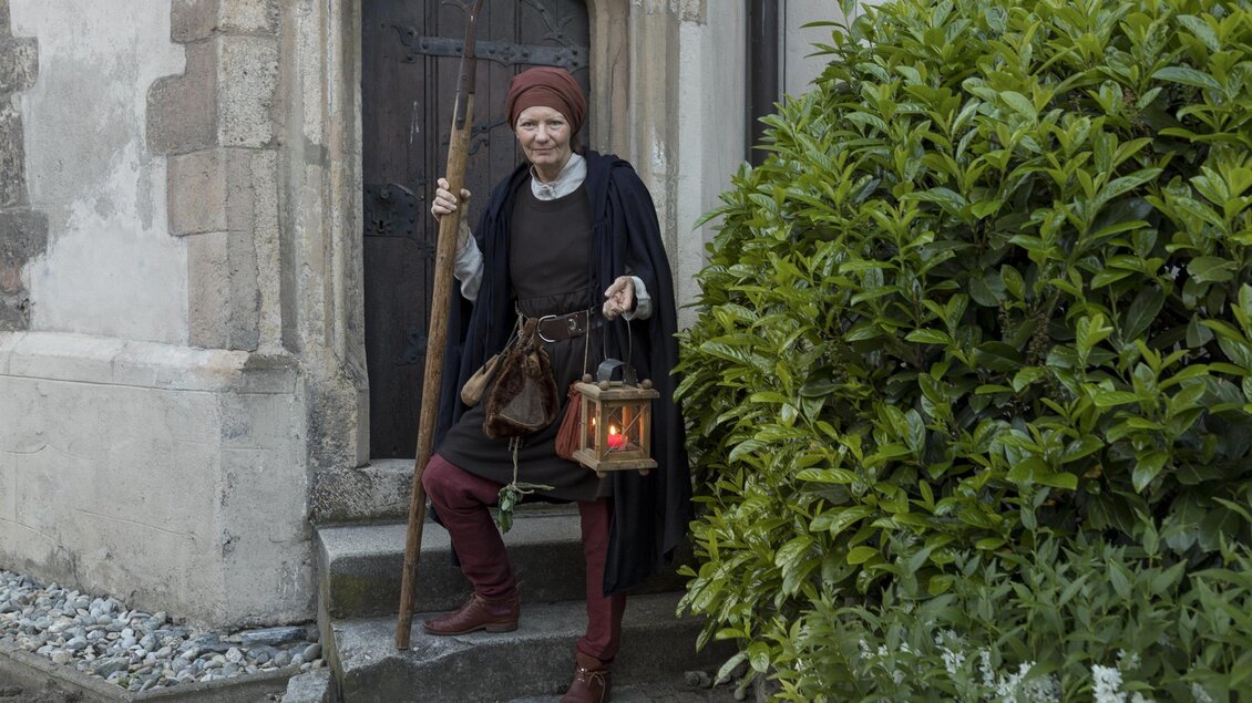 Ein mittelalterlicher Mann steht vor einer Tür und hält eine Laterne in der Hand. Er trägt traditionelle Kleidung und hat einen Gehstock dabei. | © Martin Meieregger