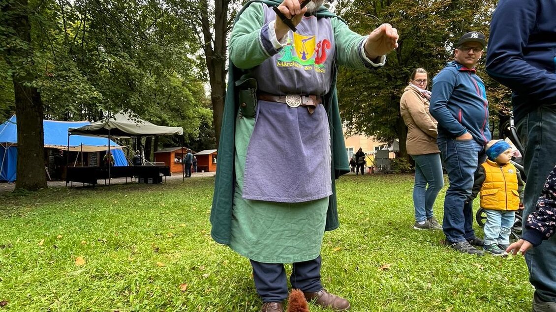 Ein Mann in historischer Kleidung steht auf einer Wiese und hält ein Spielzeug in der Hand. Im Hintergrund sind Menschen und Bäume zu sehen. | © Erlebnisregion Murtal