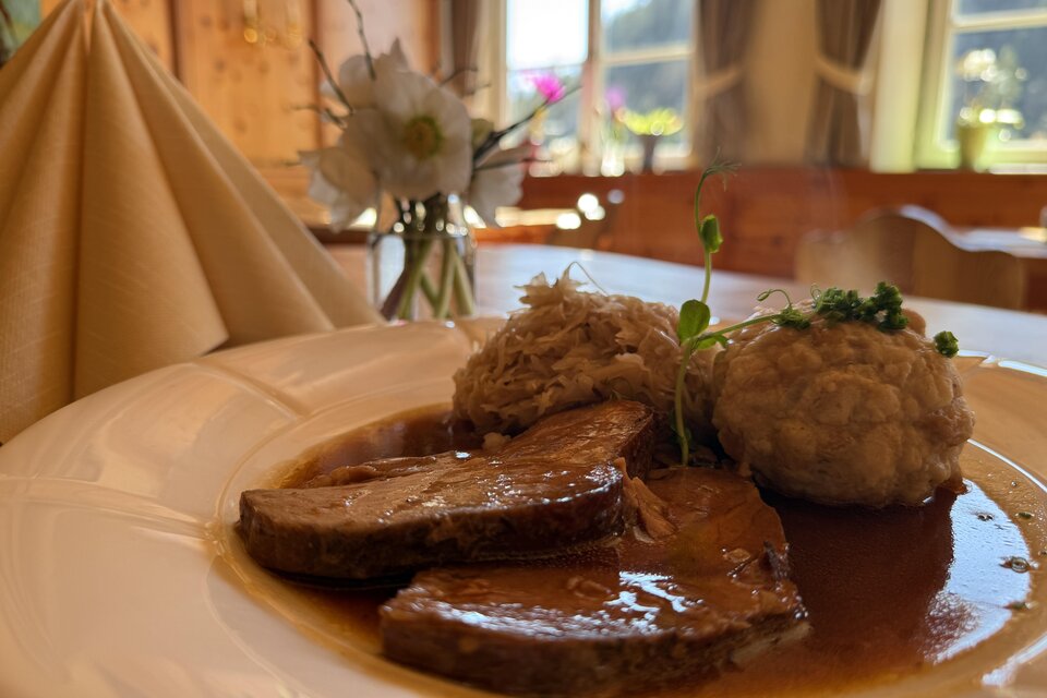 Ein leckeres Gericht mit zartem Fleisch, serviert mit Knödeln und sauerkraut. Im Hintergrund ist eine gemütliche Atmosphäre mit schönem Licht zu sehen.