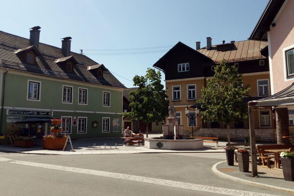 Blick auf das Kriegerdenkmal in Bad Mitterndorf mit dem Sportgeschäft Mörth und dem Restaurant S’Platzl. Im Hintergrund strahlt ein blauer, wolkenloser Himmel. | © Johanna Provatopoulos